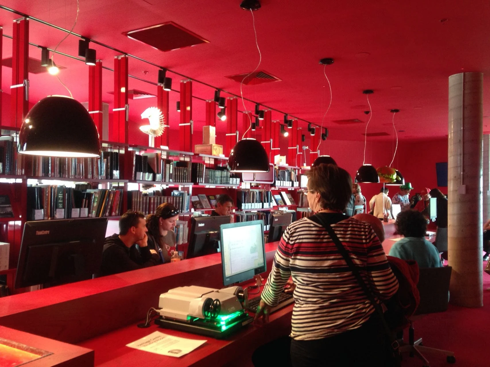 A library or bookstore with red walls and shelves filled with books. Several people are sitting or standing at a red counter with computer monitors and equipment, browsing or working. Black pendant lights hang from the ceiling, illuminating the area.
