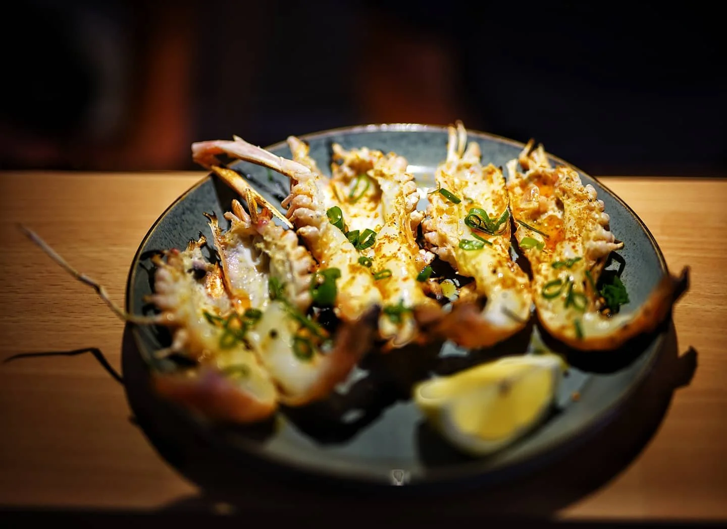 Grilled lobster halves garnished with chopped green onions, served with lemon wedges on a dark plate.
