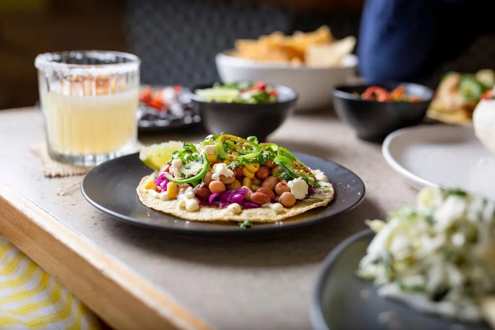 A taco on a black plate topped with beans, cheese, and green onions, with a lemon wedge on the side. In the background, there are bowls of chips, salsa, and salad, along with a glass of a light-colored drink on a wooden table.