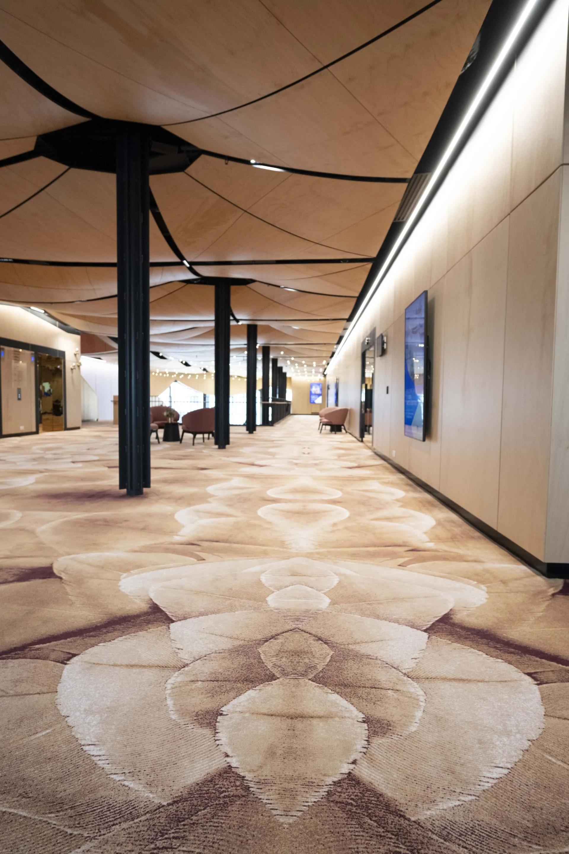 Interior of a modern hotel lobby with a patterned carpet, light wood walls, and ceiling with floating wooden panels. There are chairs and digital screens along the hallway.