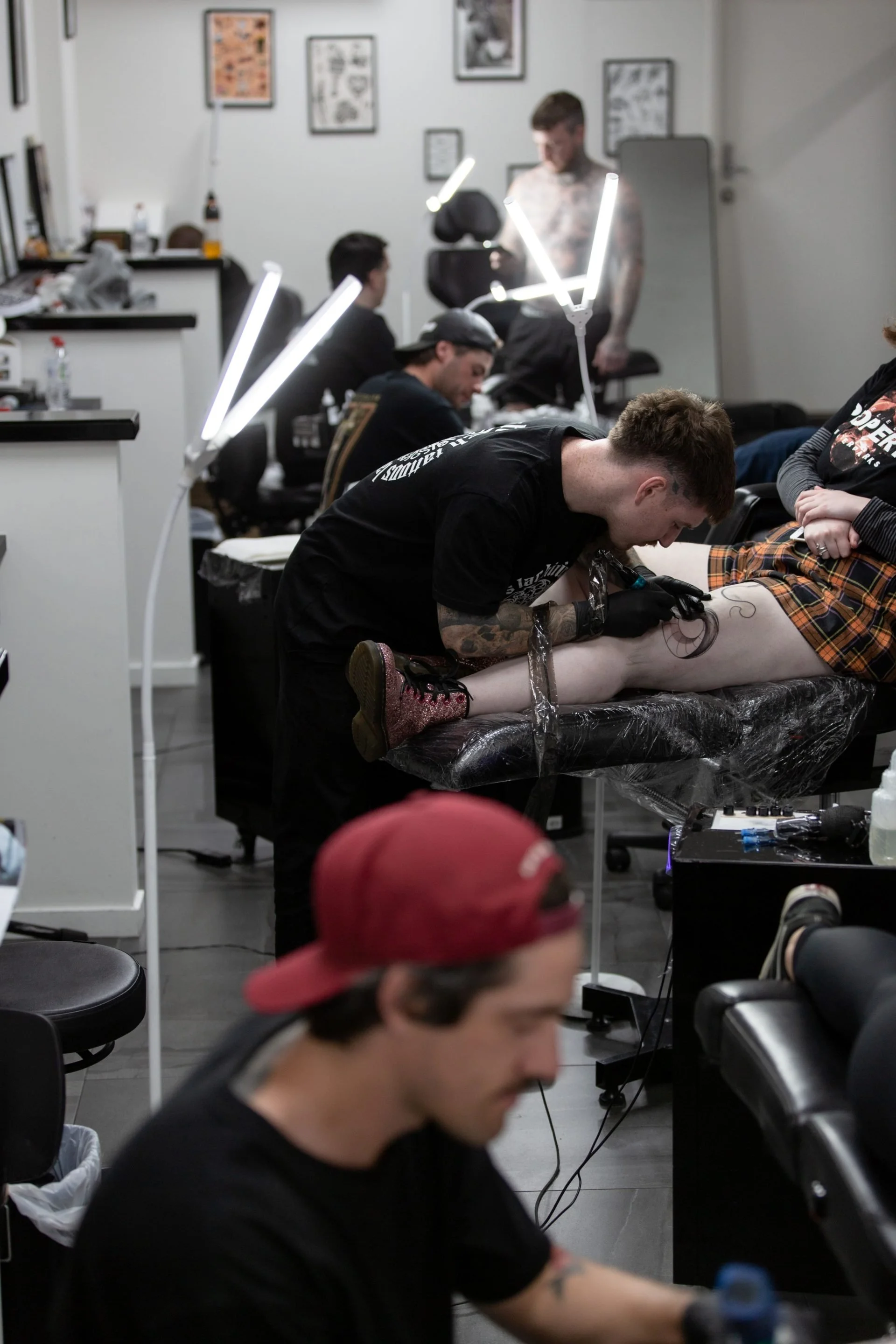 A tattoo artist working on a person's leg in a tattoo studio with other artists and clients visible in the background, and bright task lights illuminating the work area.