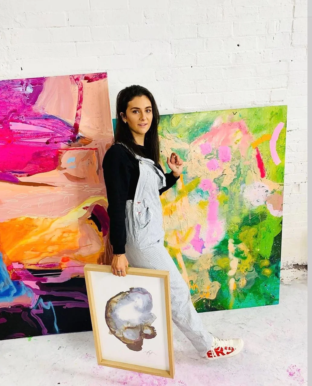Woman standing in an art studio holding a small framed watercolor painting of a person's profile in front of large colorful abstract paintings, with a white brick wall background.