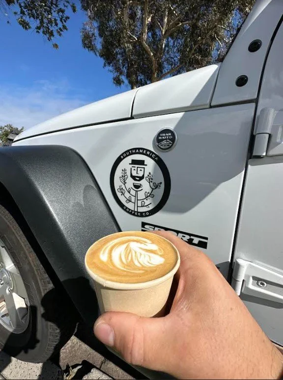 A hand holding a cup of latte with latte art in front of a white vehicle with stickers on it