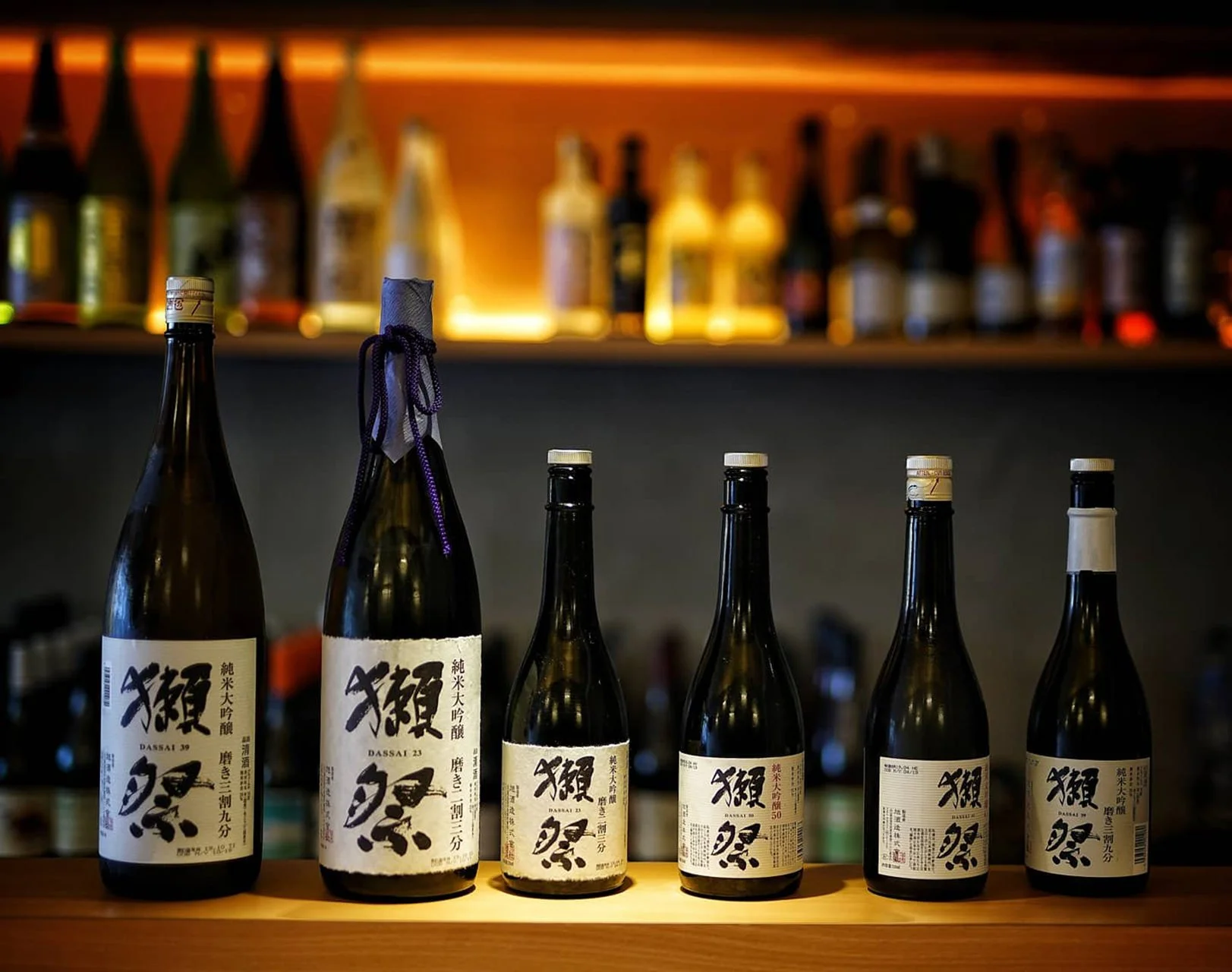 Five bottles of sake with Japanese labels on a wooden bar counter, with blurred liquor bottles and warm lighting in the background.