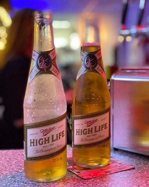 Two bottles of Miller High Life beer on a bar counter with a colorful blurred background.
