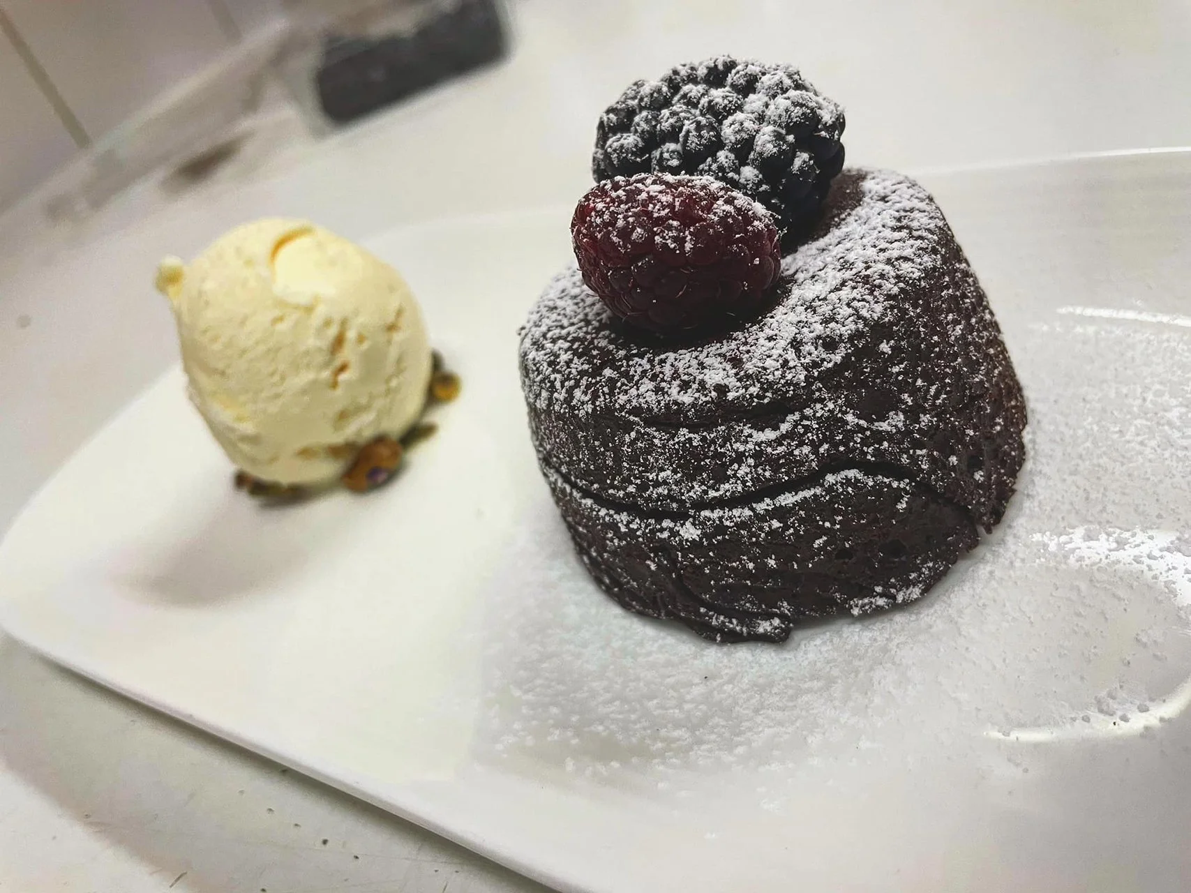 Chocolate lava cake topped with raspberries and powdered sugar, served with a scoop of vanilla ice cream on a white plate.