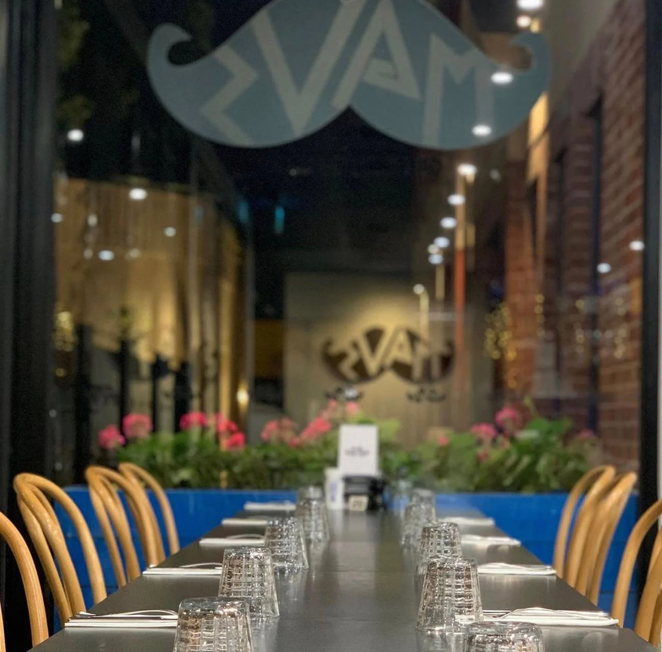 Empty restaurant table set with glasses and utensils at night, with pink flowers and a decorative wall sign in the background.