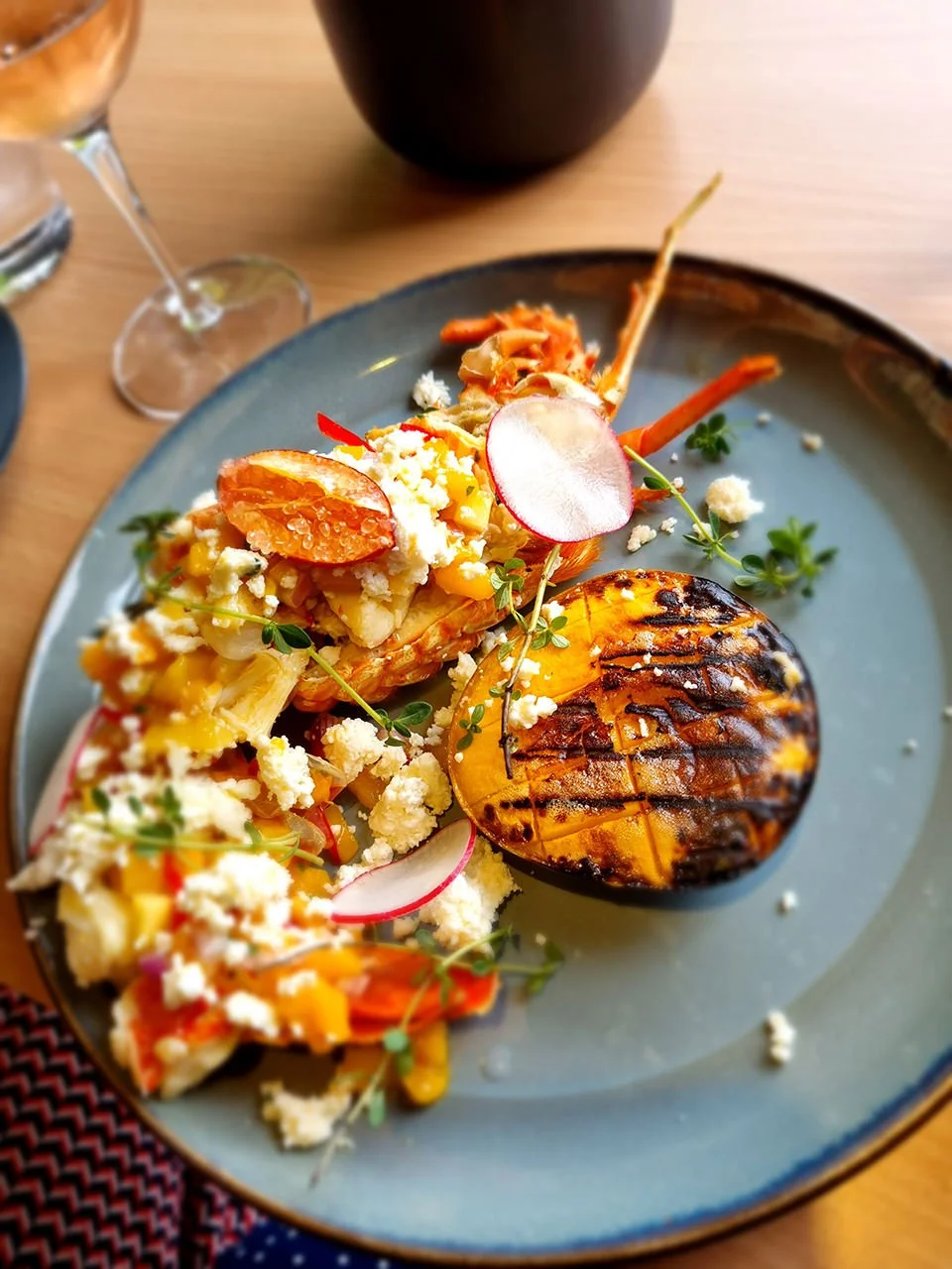 Grilled lobster tail served with vegetables, radish slices, crumbled cheese, garnished with microgreens, on a blue plate.