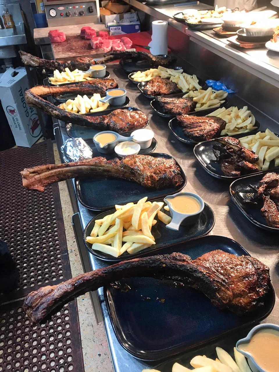 A row of cooked lamb chops with long bones, French fries, and small bowls of sauce on black trays in a restaurant kitchen.