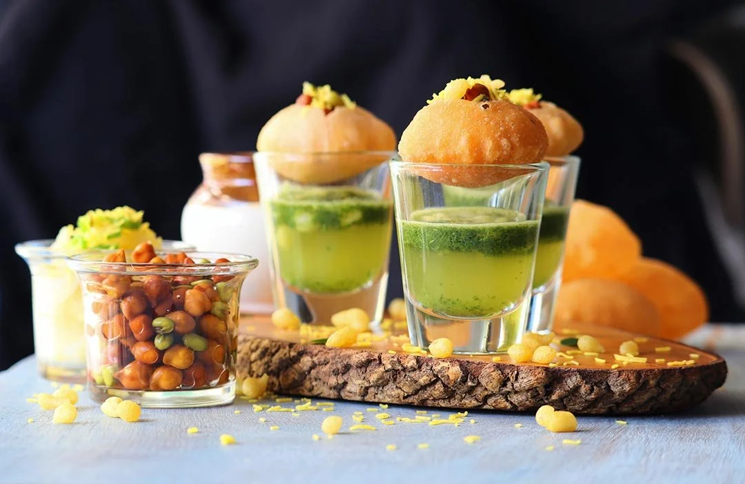 Assorted Indian sweets and snacks, including falooda, chickpeas, and ladoos, served on a wooden board and glass cups.