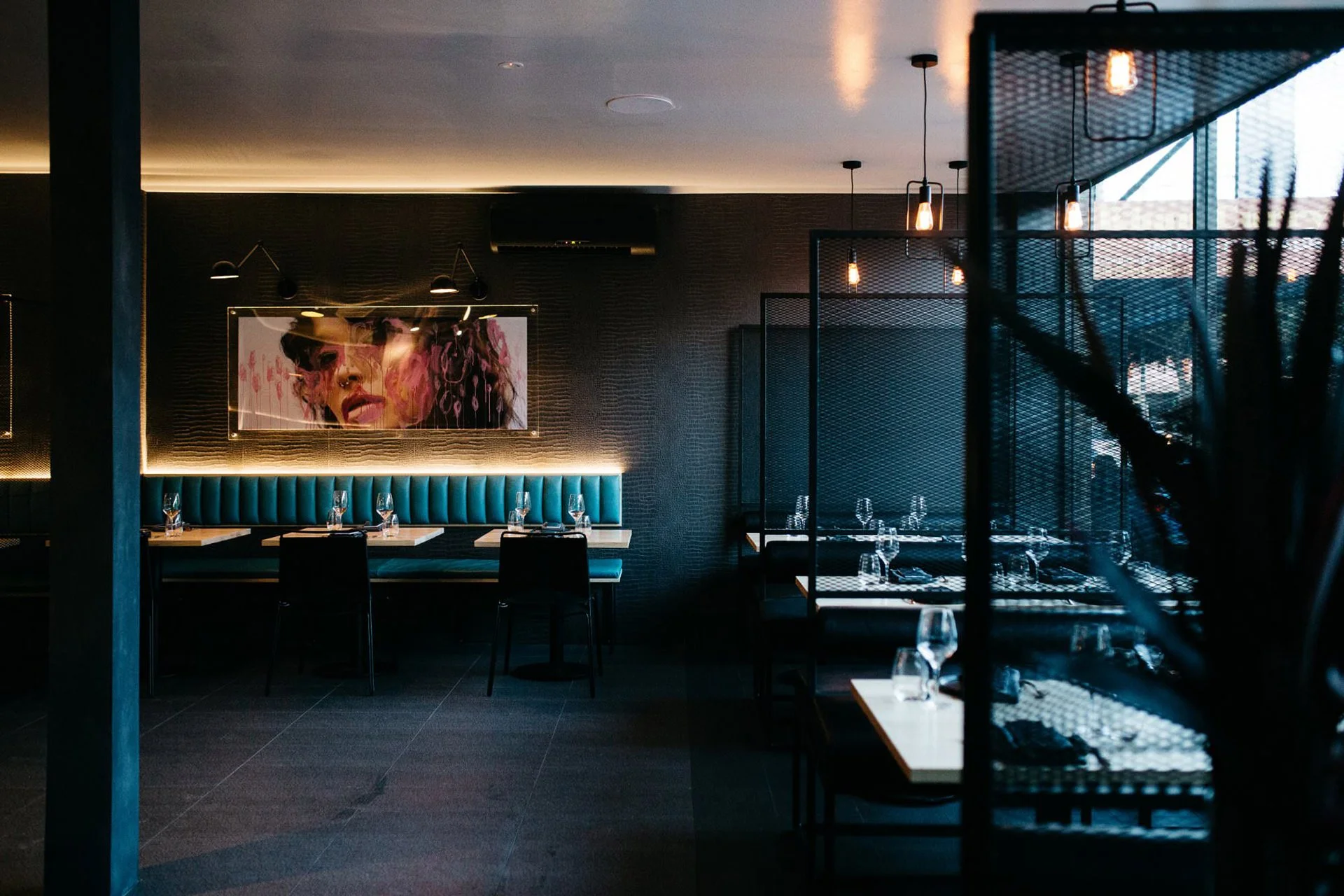 Modern restaurant dining area with teal booth seating, black chairs, and tables set with glassware. Dark walls decorated with a large portrait of a woman with sunglasses. Warm lighting and a partially visible window with outdoor light.