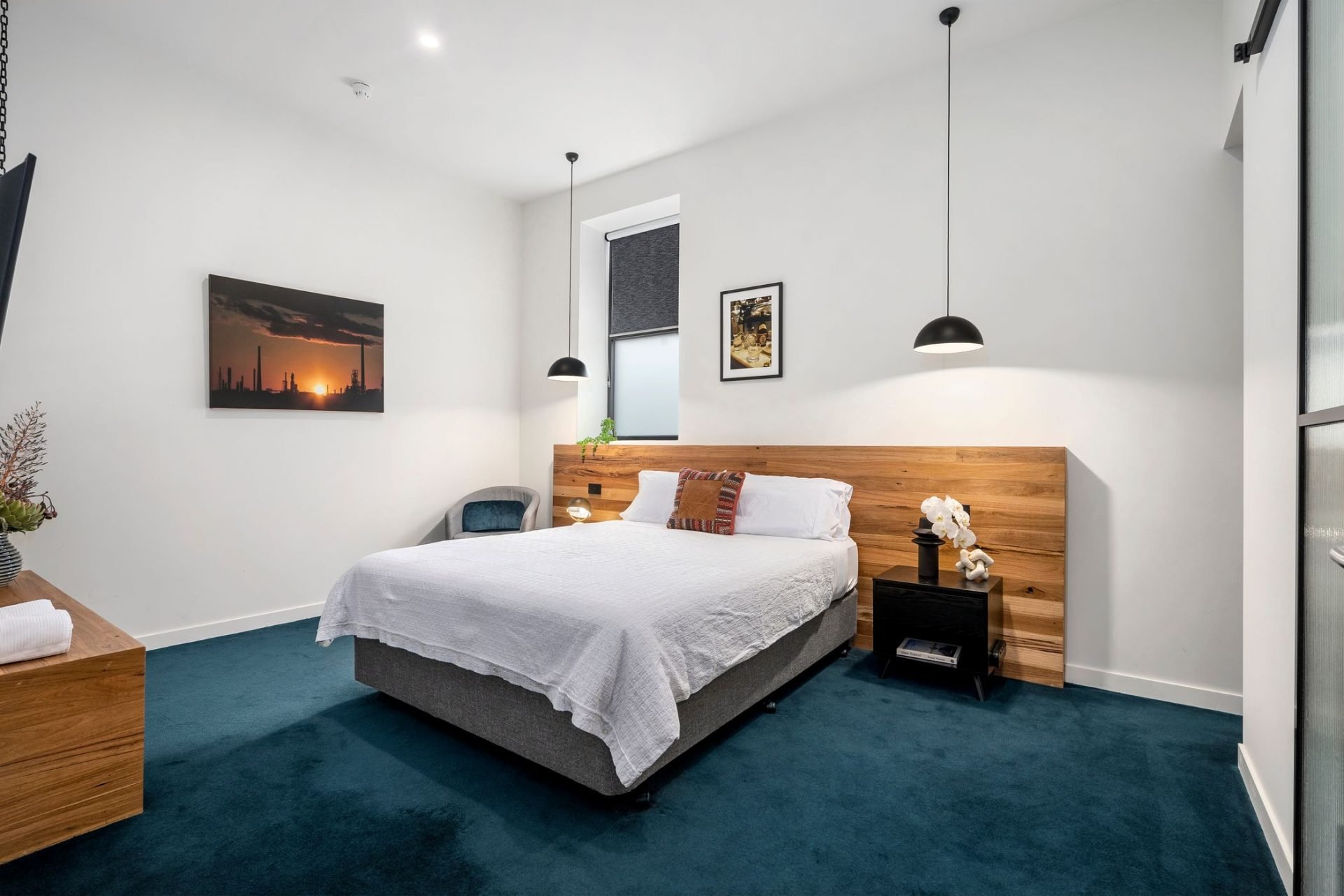 Modern bedroom with white walls, a large bed with white linens and pillows, wooden headboard, dark blue carpet, black pendant lights, framed artwork, and a window with black blinds.