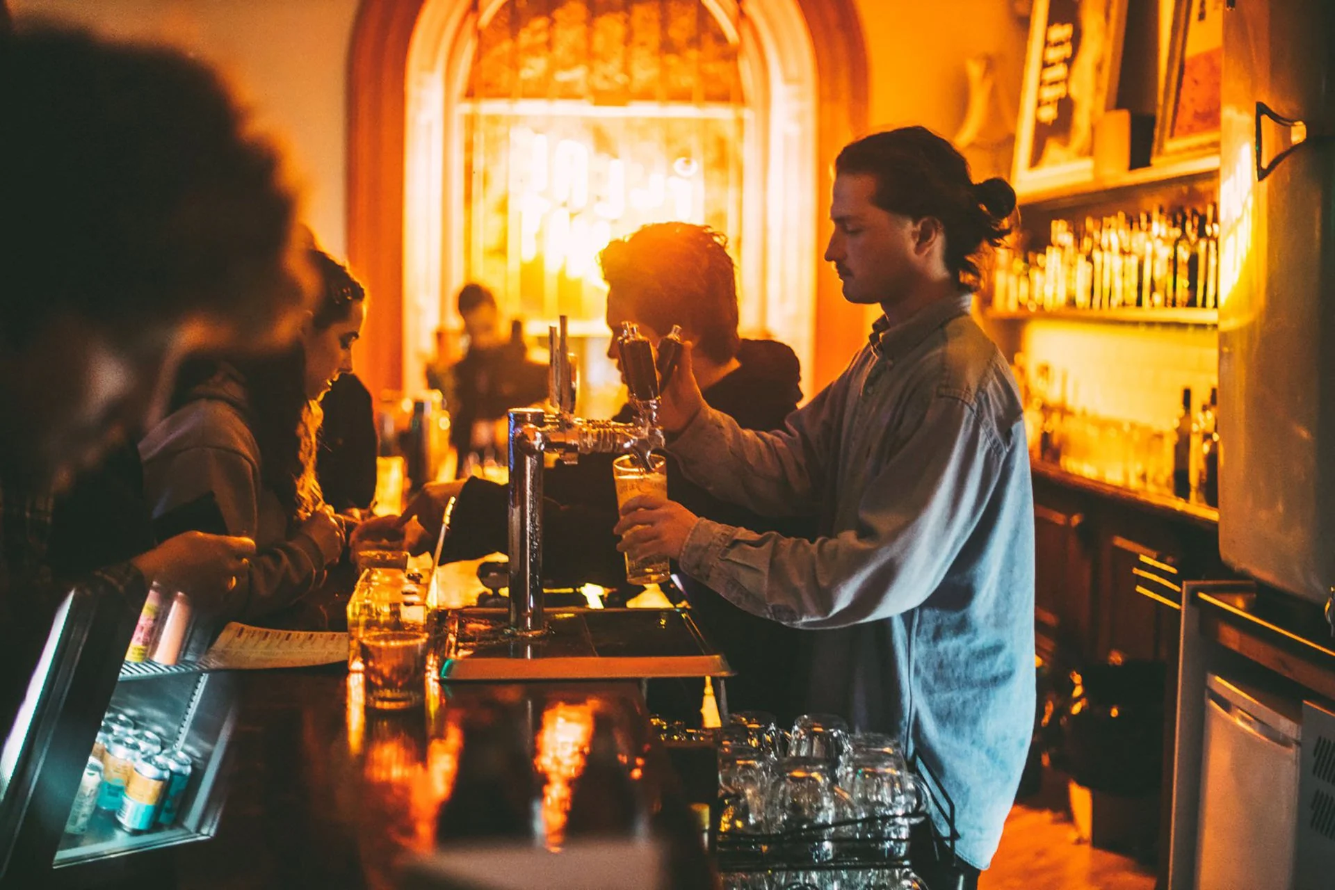 Bar scene with bartender pouring beer and customers ordering drinks in dim, warm lighting.
