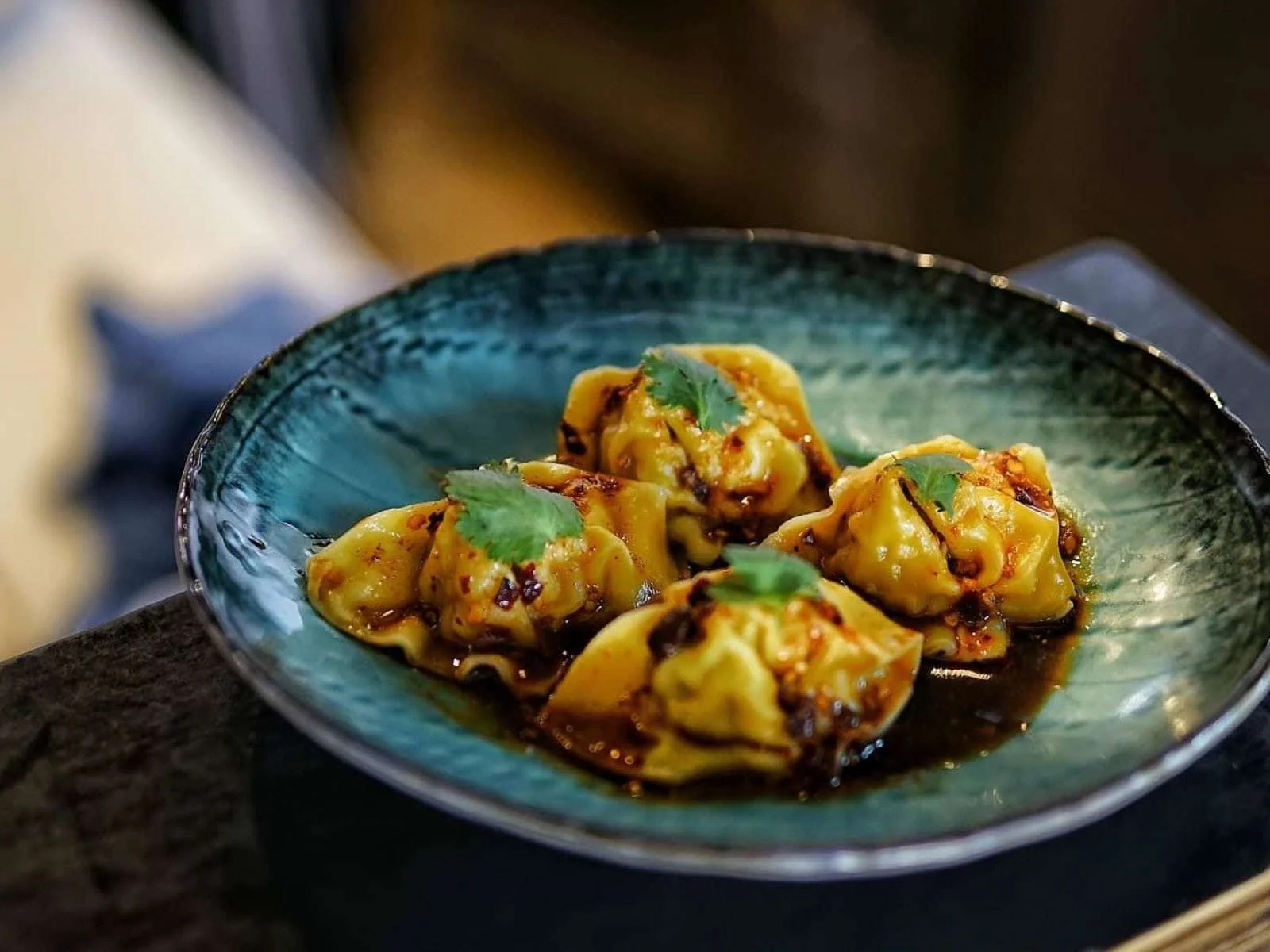 Close-up of a blue ceramic plate with five Asian-style dumplings garnished with cilantro leaves, sitting in a dark sauce.