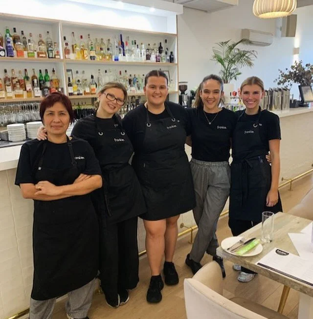 Group of five women in black aprons standing together in a restaurant or bar, smiling, with a well-stocked bar in the background.