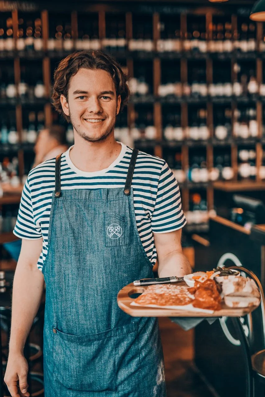 A young man with wavy brown hair, smiling, wearing a striped t-shirt and a denim apron, serving food on a wooden platter in a restaurant with wine bottles on shelves in the background.
