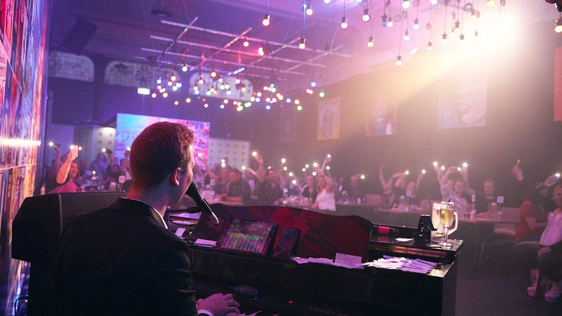 A pianist performing on stage at a nightclub, with an audience in the background cheering and holding up phones. The club has colorful lighting and decorations.