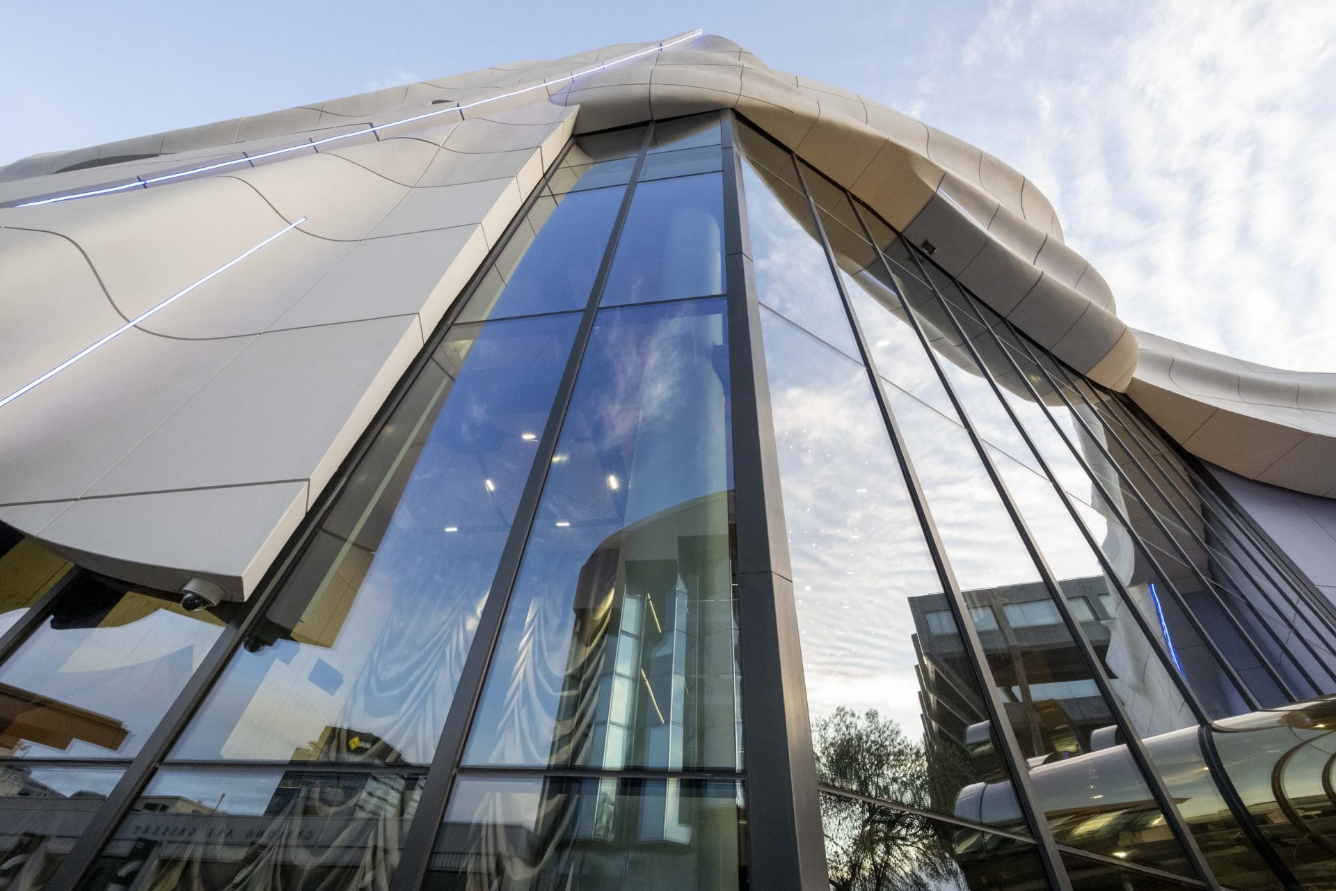 Low-angle view of a modern architectural building with large glass windows and smooth metallic exterior, reflecting the cloudy sky.