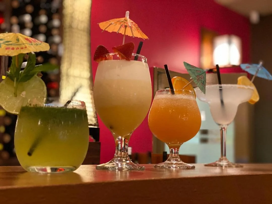Four colorful cocktails with small umbrellas and garnishes on a wooden bar in a cozy, warmly lit setting.