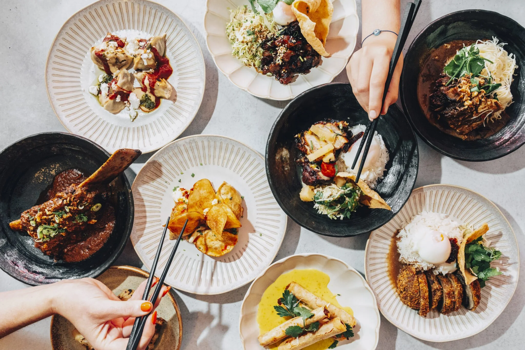 A top-down view of a variety of Asian dishes on plates and bowls, including rice, noodles, curry, and fried appetizers, with some chopsticks.