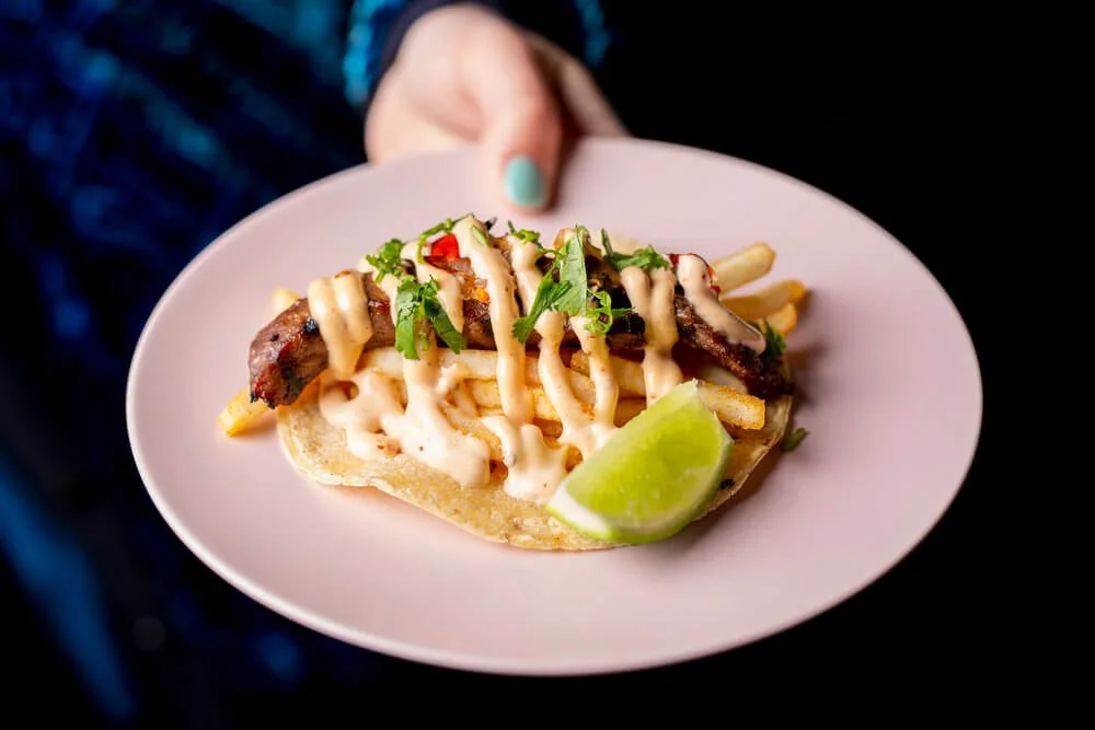 A plate of gourmet taco with grilled meat, topped with mayonnaise sauce, chopped green herbs, sliced red pepper, and a lime wedge.