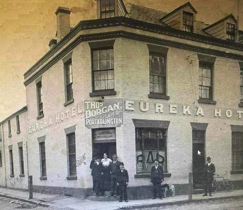 Black and white photo of a building labeled 'Eureka Hotel' with a sign for a Dob Cani (hot dogs) stand. Five men and a woman are standing in front, some with hats. One man is standing with a dog, and another with a bicycle.