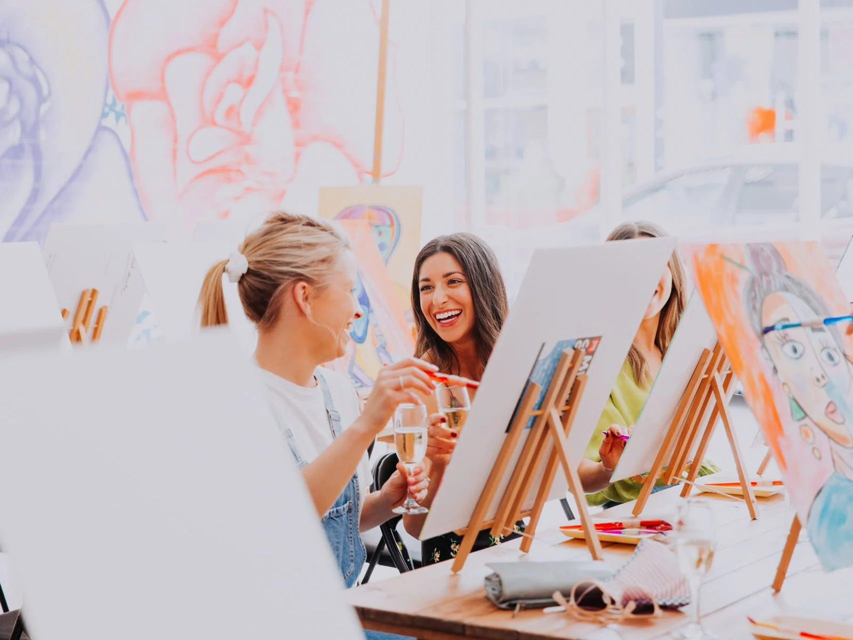 A group of women participating in a painting class, smiling and chatting while painting on canvases. The setting is bright and colorful, with art supplies on the table and artwork on the walls.
