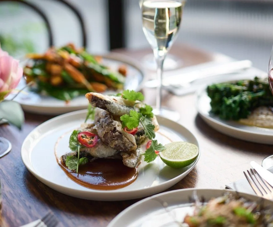 Fried fish served with lime and garnished with cilantro and red chili slices on a white plate, with a brown sauce underneath, on a wooden table with other plated dishes and a glass of white wine in the background.
