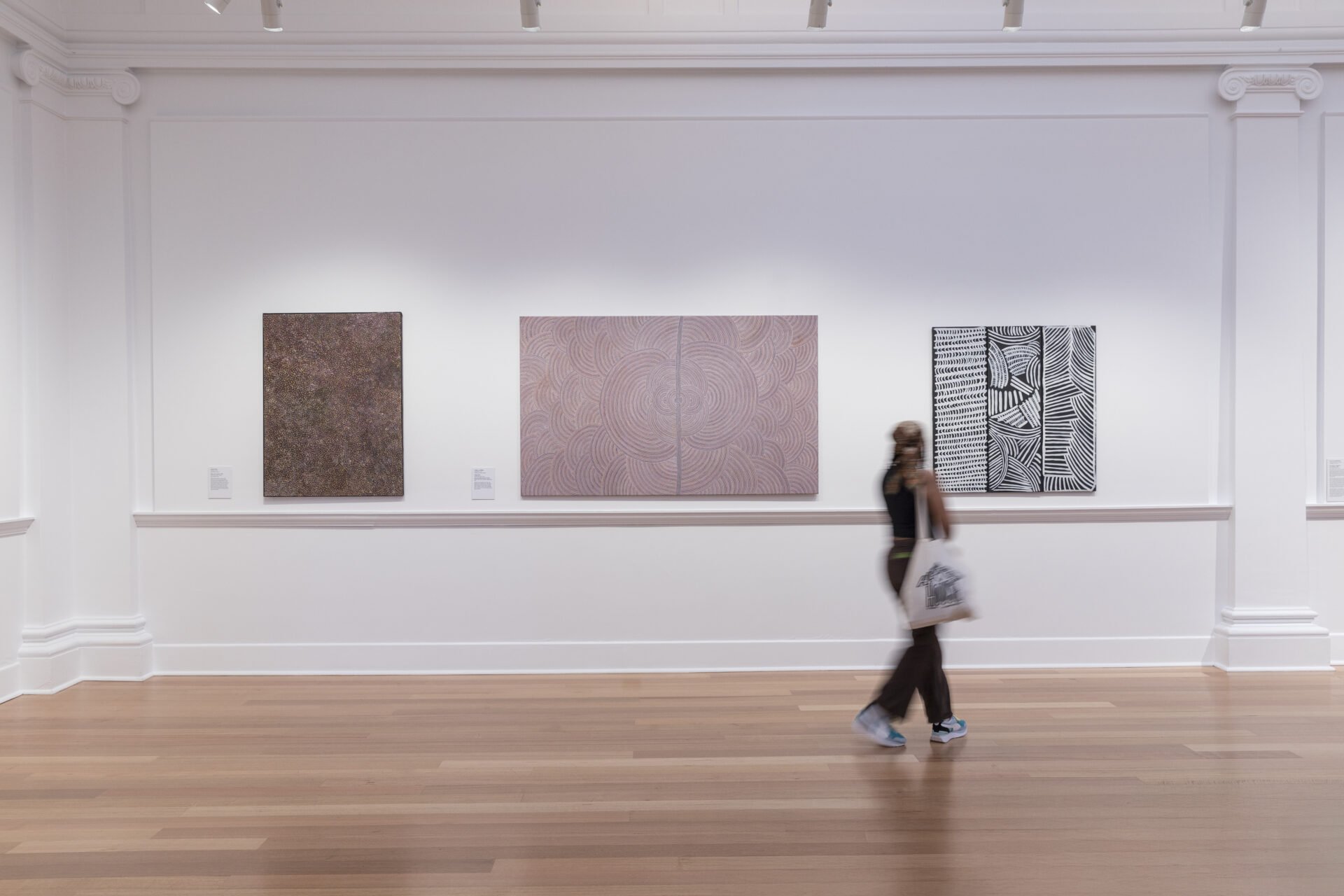 A woman walking in an art gallery with three abstract paintings on a white wall behind her, hardwood floor in the foreground.