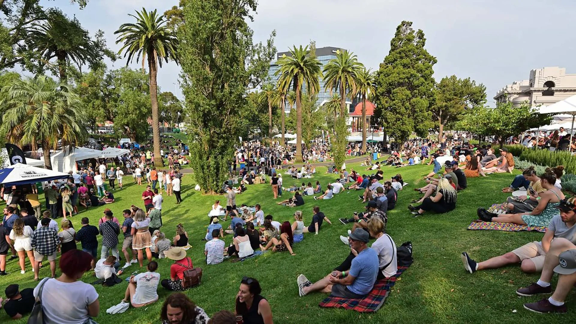 A large outdoor gathering in a park with many people sitting on the grass, some on blankets, and some standing. Tall palm trees and lush green trees surround the area, with a crowd in the background near vendor tents and a cityscape with buildings.