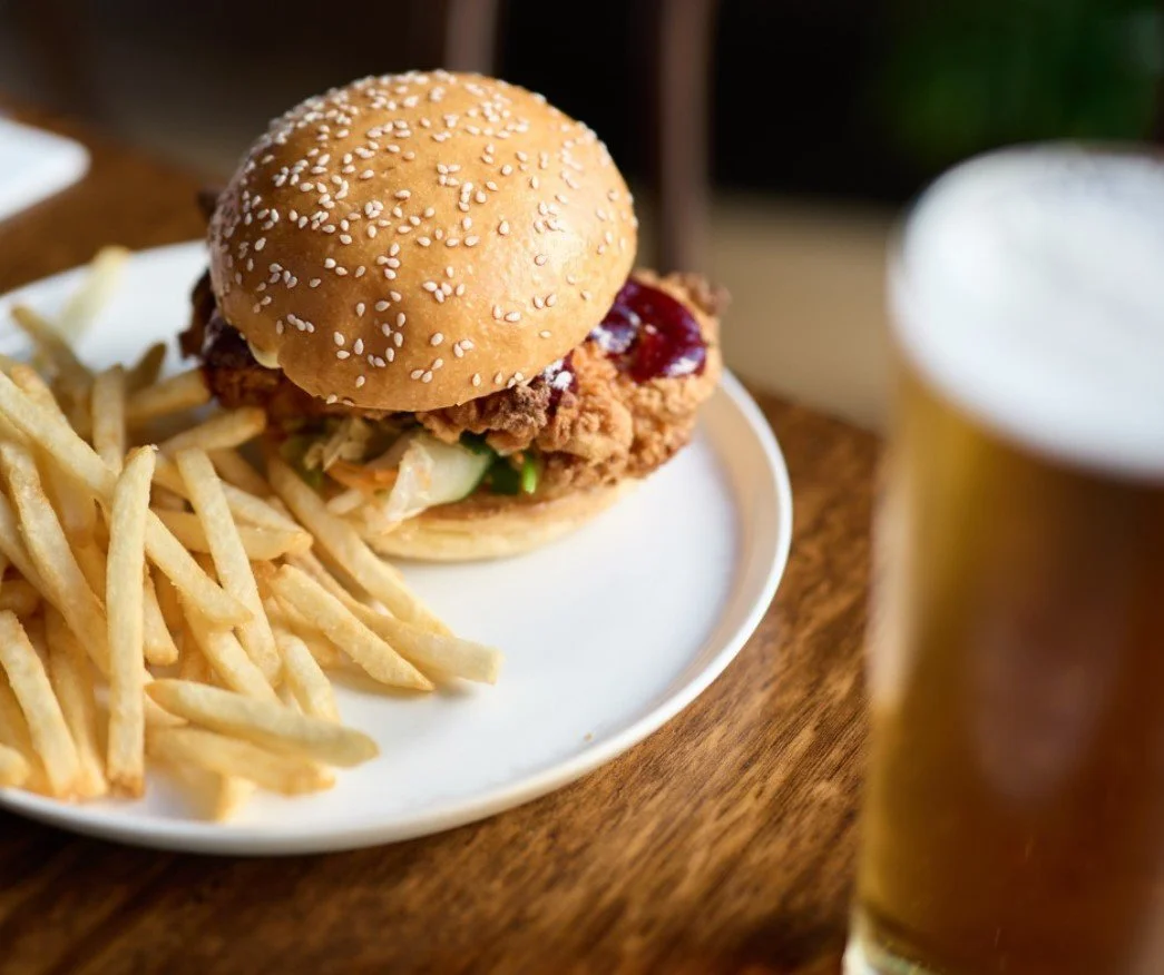 Fried chicken sandwich with lettuce and sauce on a toasted bun, served with French fries on a white plate, and a glass of beer in the background.