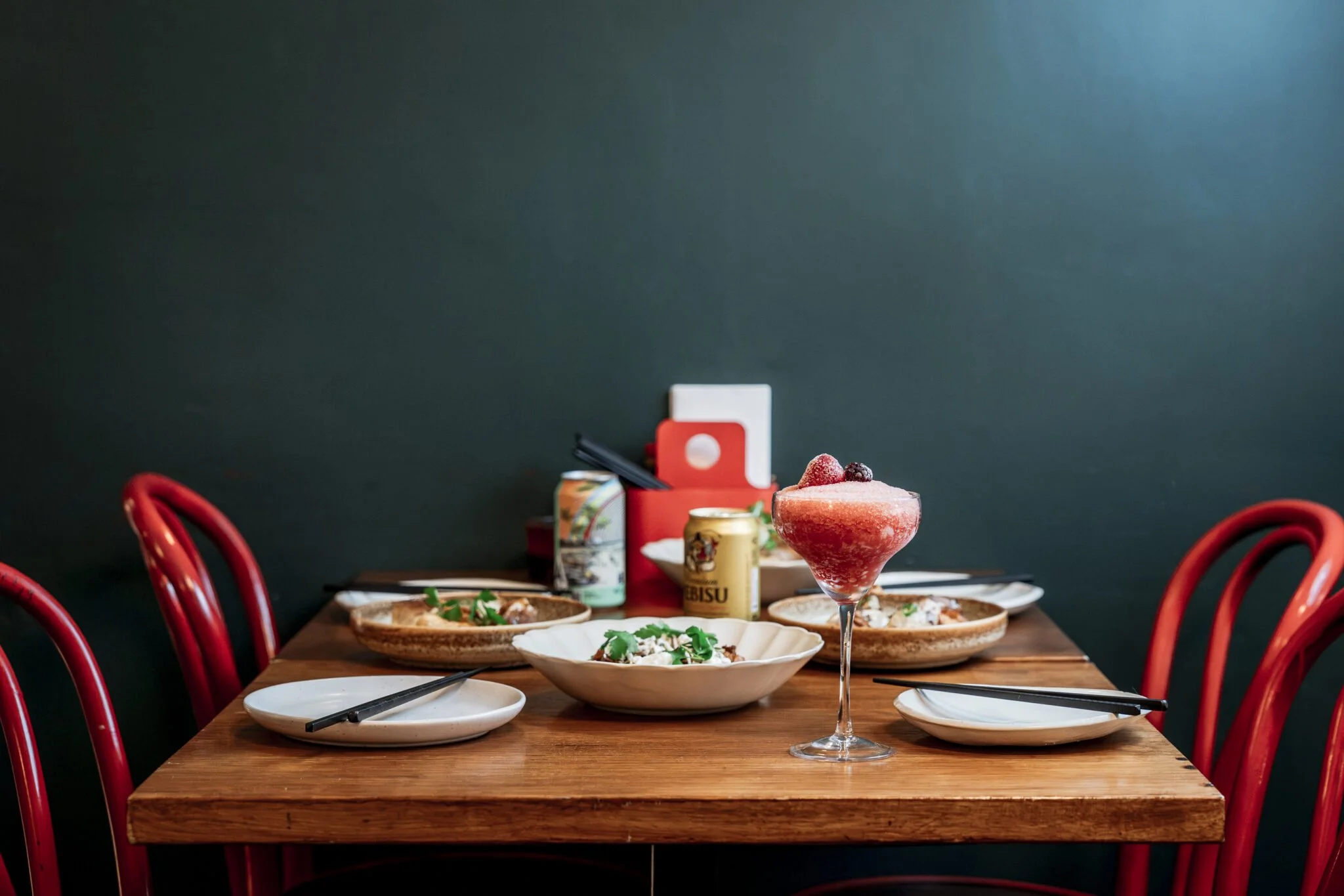 A restaurant table set with plates of food, a red cocktail garnished with berries, and canned beverages, with red chairs and a dark wall in the background.