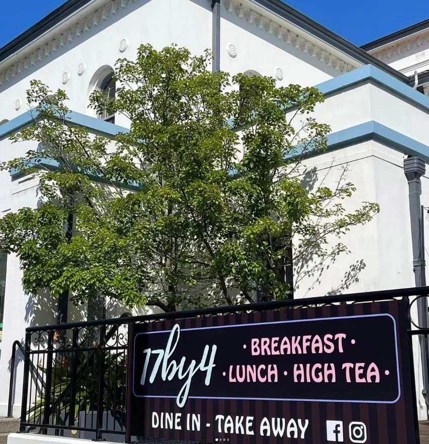 A sign for a cafe named "77buzz" advertising breakfast, lunch, and high tea, offering dine-in and take away options, placed in front of a white building with blue accents and a tree.