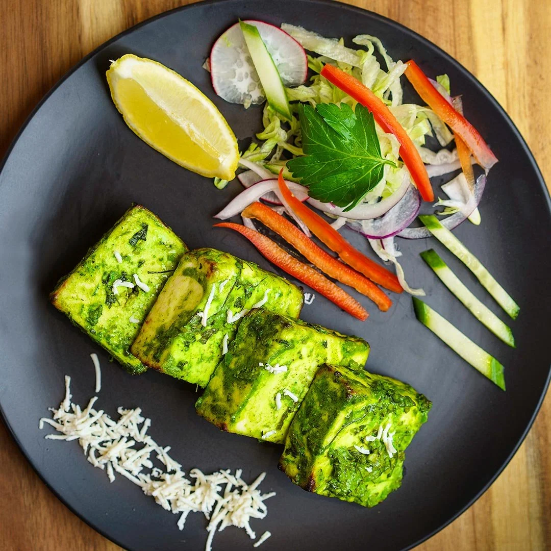 Grilled paneer cheese with green sauce on a black plate, side salad with radish, cucumber, red bell pepper, onion, and a lemon wedge.