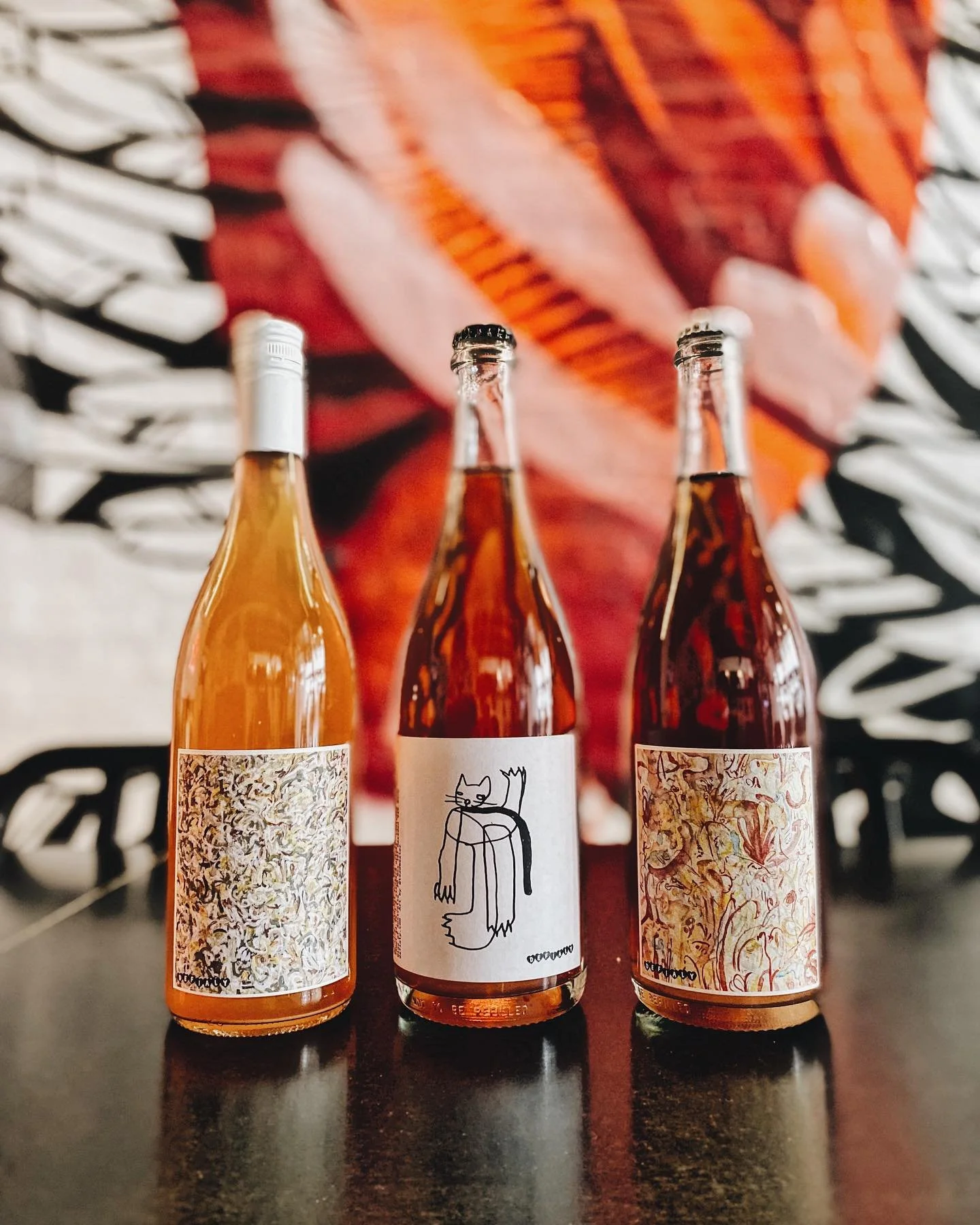 Three bottles of wine or spirits with artistic labels on a dark table, with a blurry colorful abstract artwork in the background.