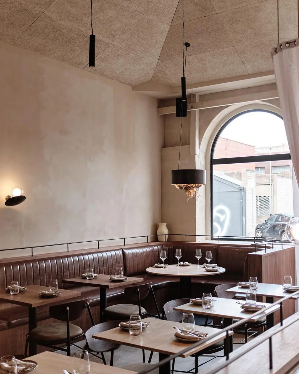 Interior of a restaurant with wooden tables set with plates, glasses, and napkins, a curved leather booth along the wall, and large arched window letting in daylight.