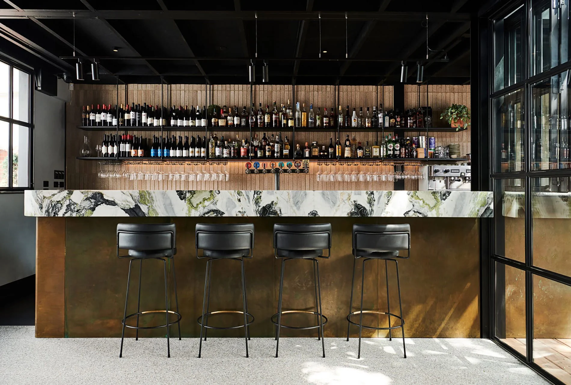 Modern bar with a white marble counter, four black bar stools, and shelves with various bottles of alcohol and glassware behind it, in a well-lit room with large windows and black ceiling.