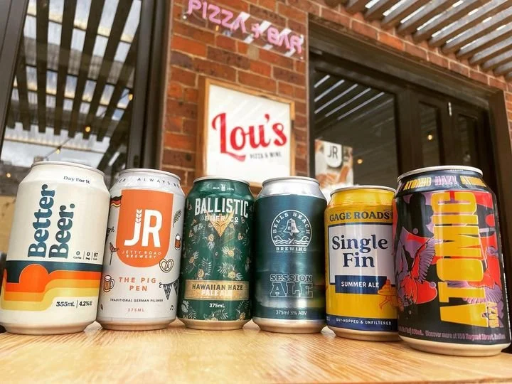 Six cans of different craft beers on a wooden table outside a brick building with a sign for Lou's and a neon sign, in front of a window and a door.