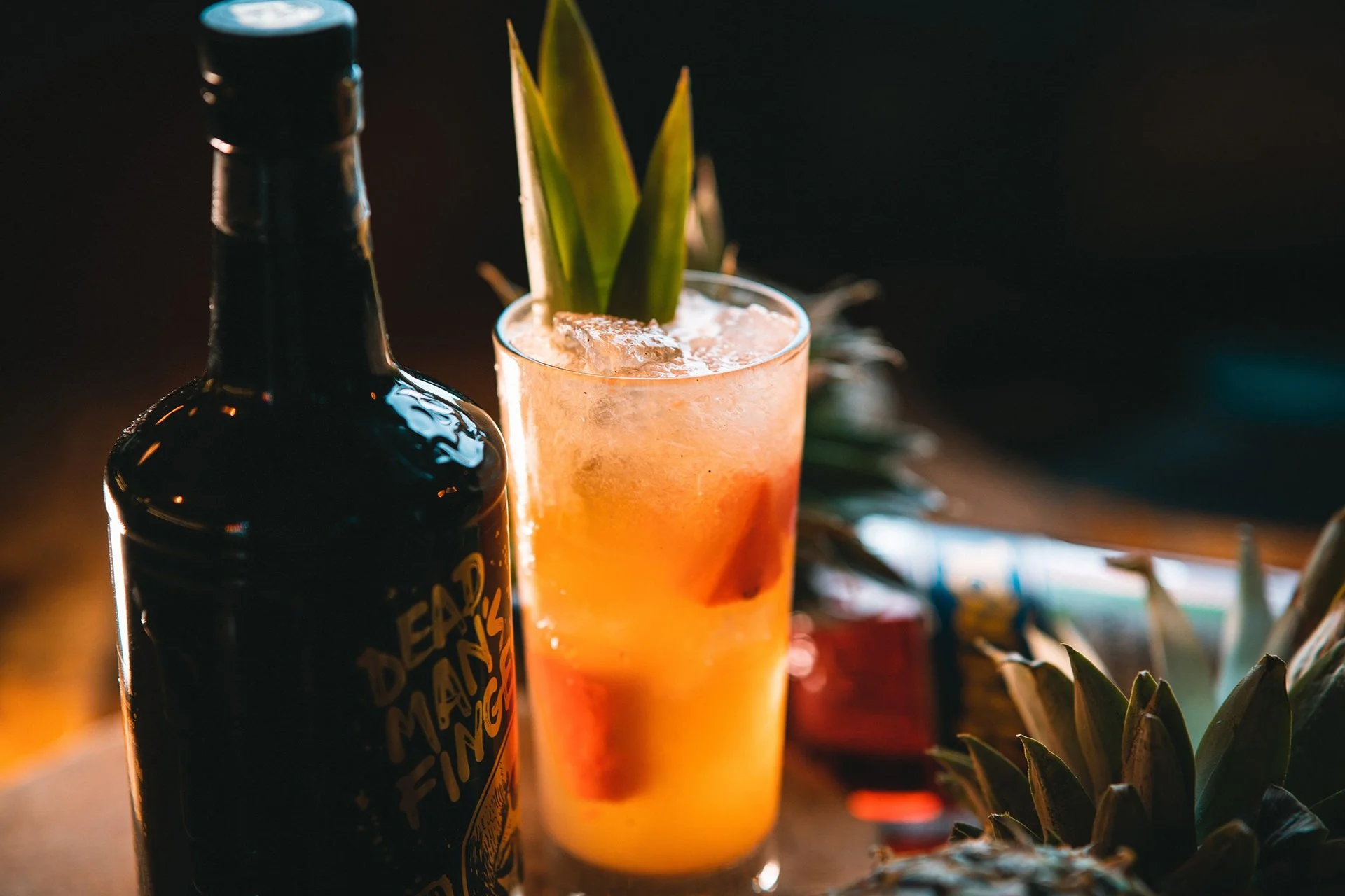 A black bottle with colorful text next to a tropical cocktail in a tall glass with pineapple leaves garnish, with pineapples and other tropical decor in the background.