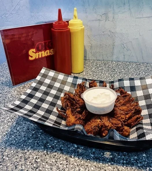 Fried chicken wings with a side of white dipping sauce on a checkered paper-lined tray, ketchup and mustard bottles behind, and a red menu or sign in the background.