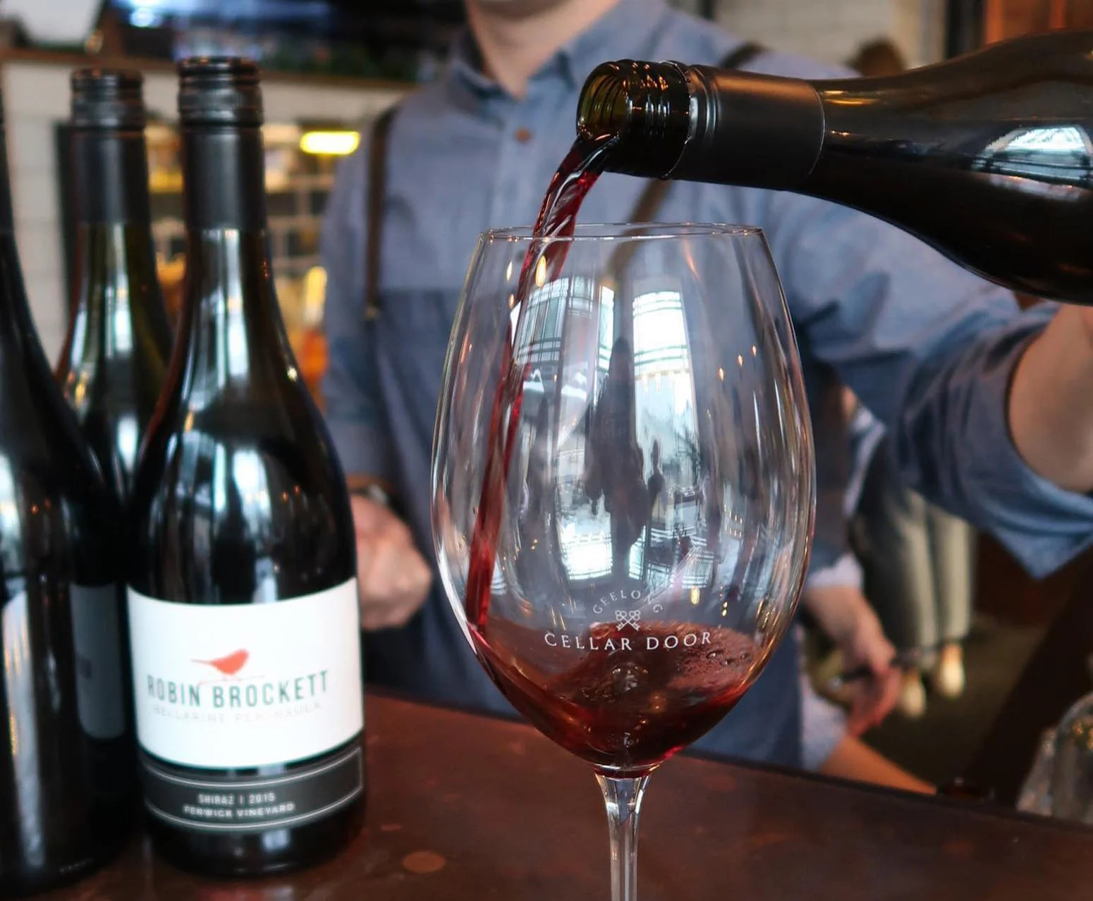 Red wine being poured from a bottle into a wine glass at a winery or tasting room.