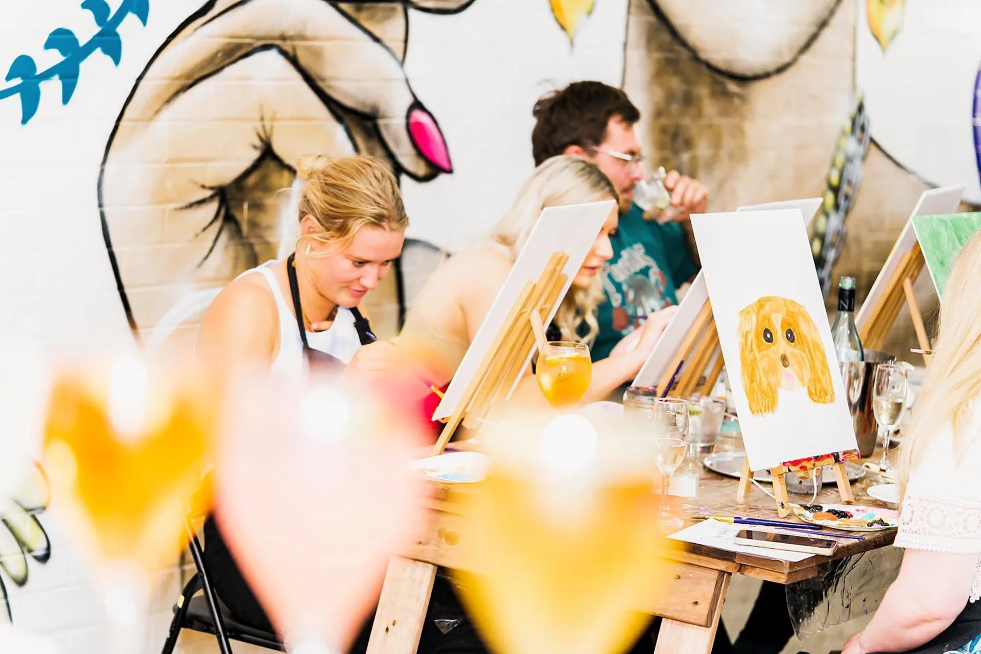 People seated at a table painting on canvases during a painting class, with colorful artwork and art supplies visible.
