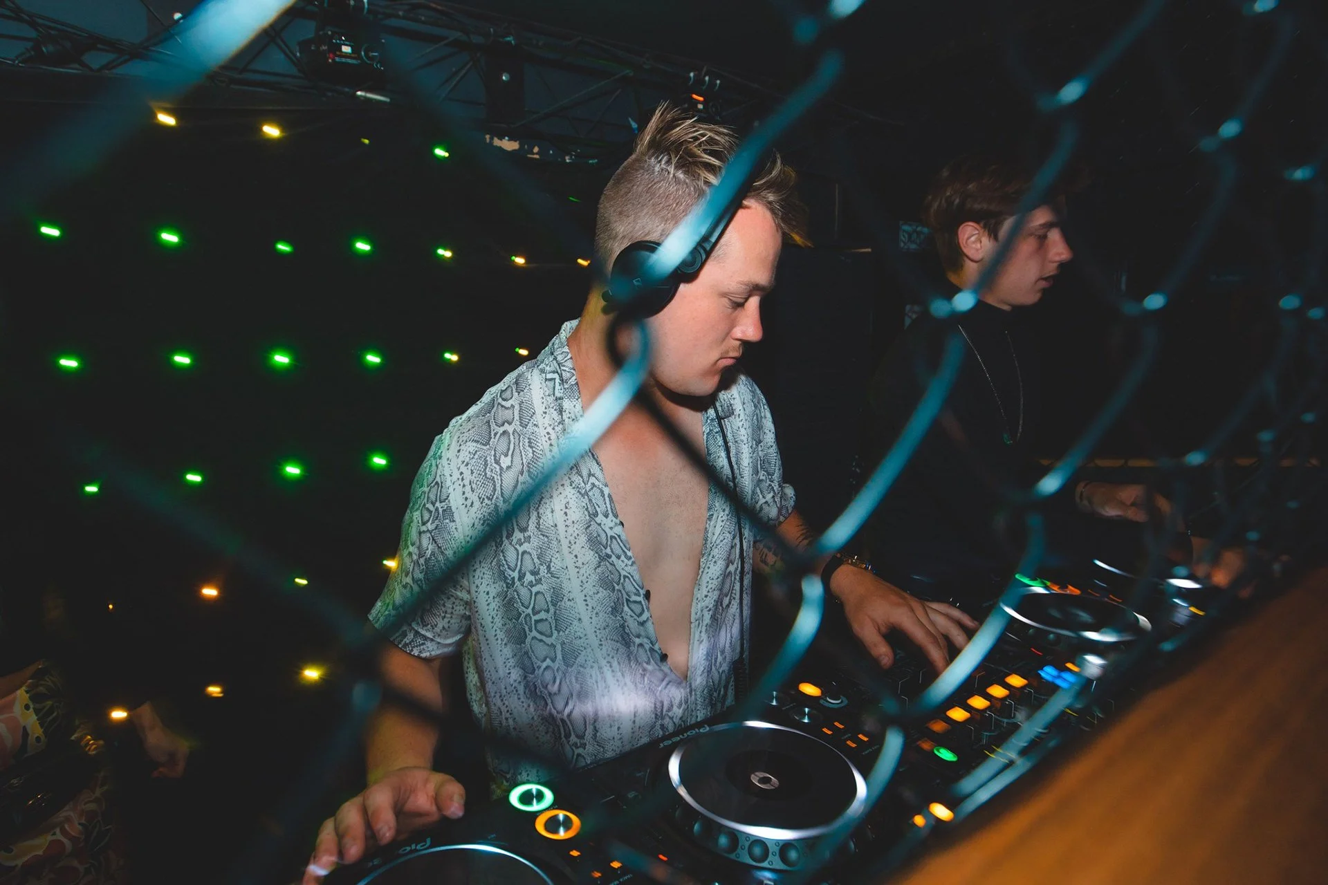 Two DJs wearing headphones and performing on a DJ console in a dark club with green, yellow, and orange lights visible in the background.