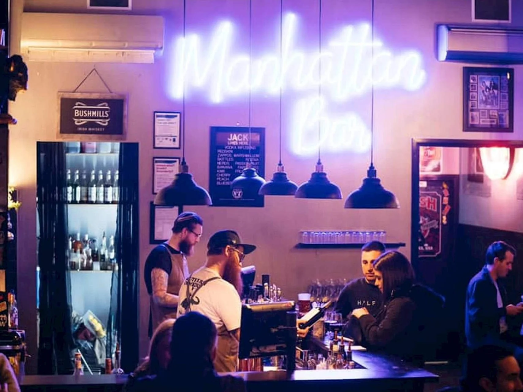 A bar with neon sign reading 'Manhattan Bar'. Several patrons are seated or standing, with bartenders behind the counter preparing drinks. The setting has dim lighting and a casual atmosphere.
