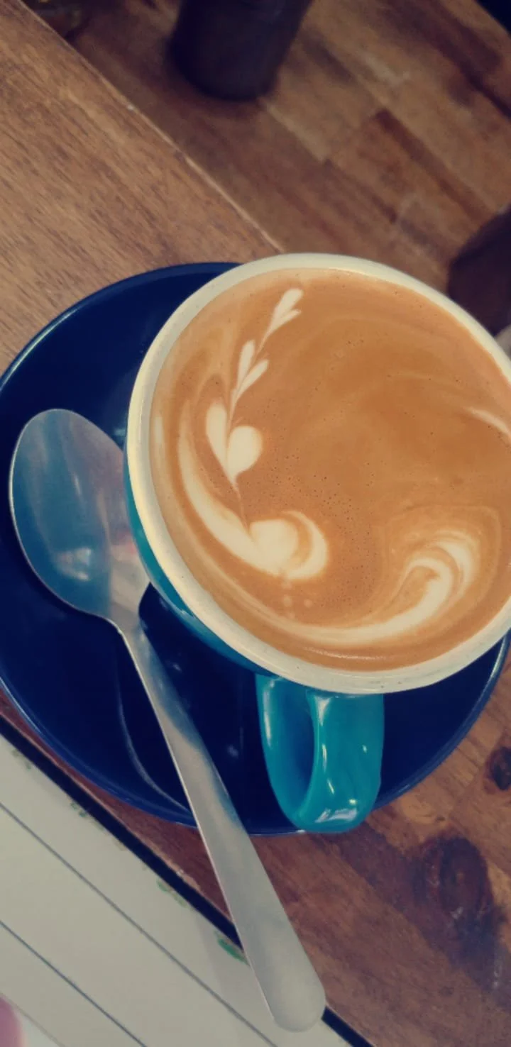 A cup of latte with latte art on top, placed on a dark blue saucer with a silver spoon beside it on a wooden table.