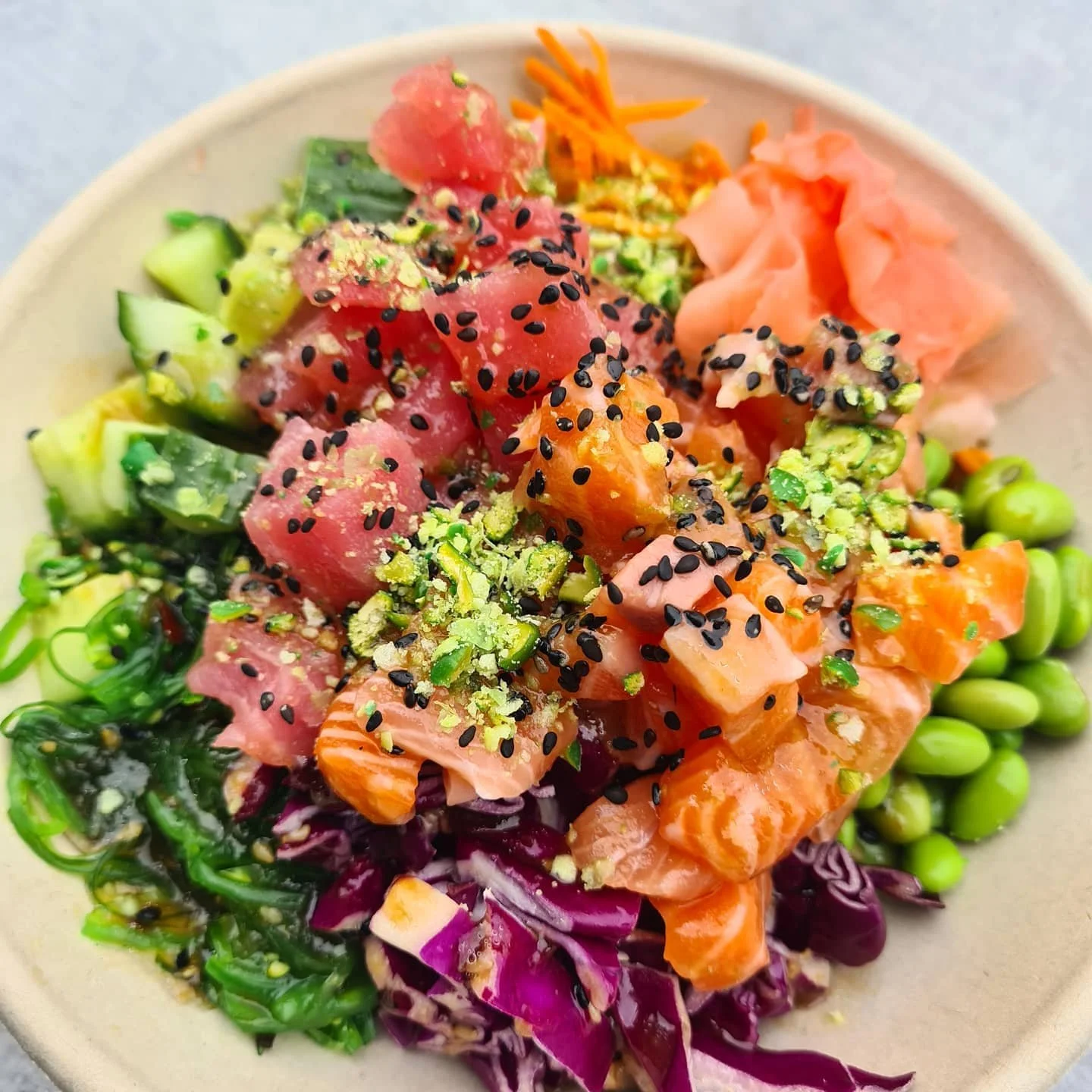A bowl of poke salad with fresh fish, avocado, seaweed, edamame, shredded purple cabbage, carrot, and green onions garnished with black sesame seeds and chopped pistachios.
