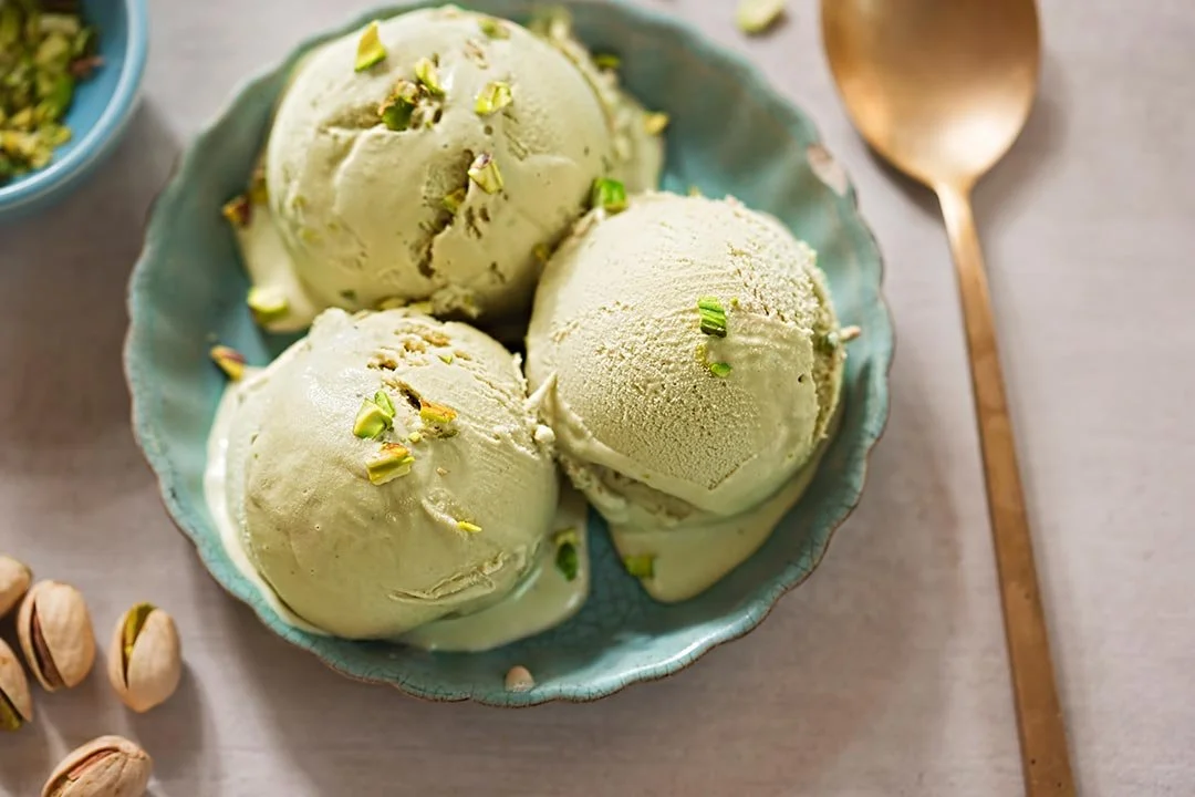 Three scoops of pistachio ice cream in a blue bowl, garnished with chopped pistachios, on a light-colored table with a gold spoon nearby.