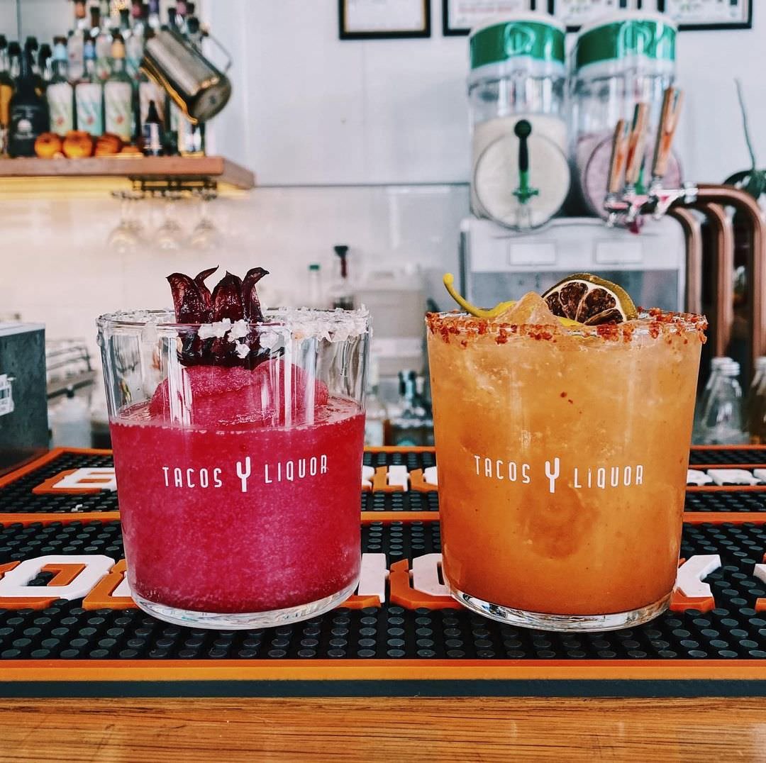 Two colorful cocktails with salted rims on a bar counter, one pink with beet garnish and the other orange with lemon and lime slices, at Tacos y Liquor.