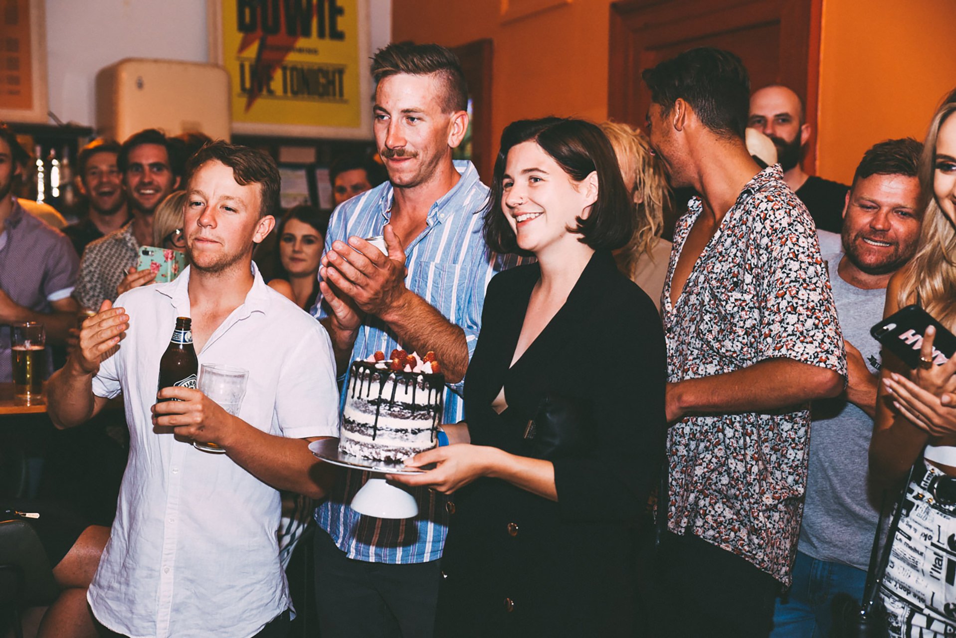 People celebrating a birthday, with a woman holding a cake with strawberries, chocolate drizzle, and whipped cream, surrounded by friends clapping and smiling.