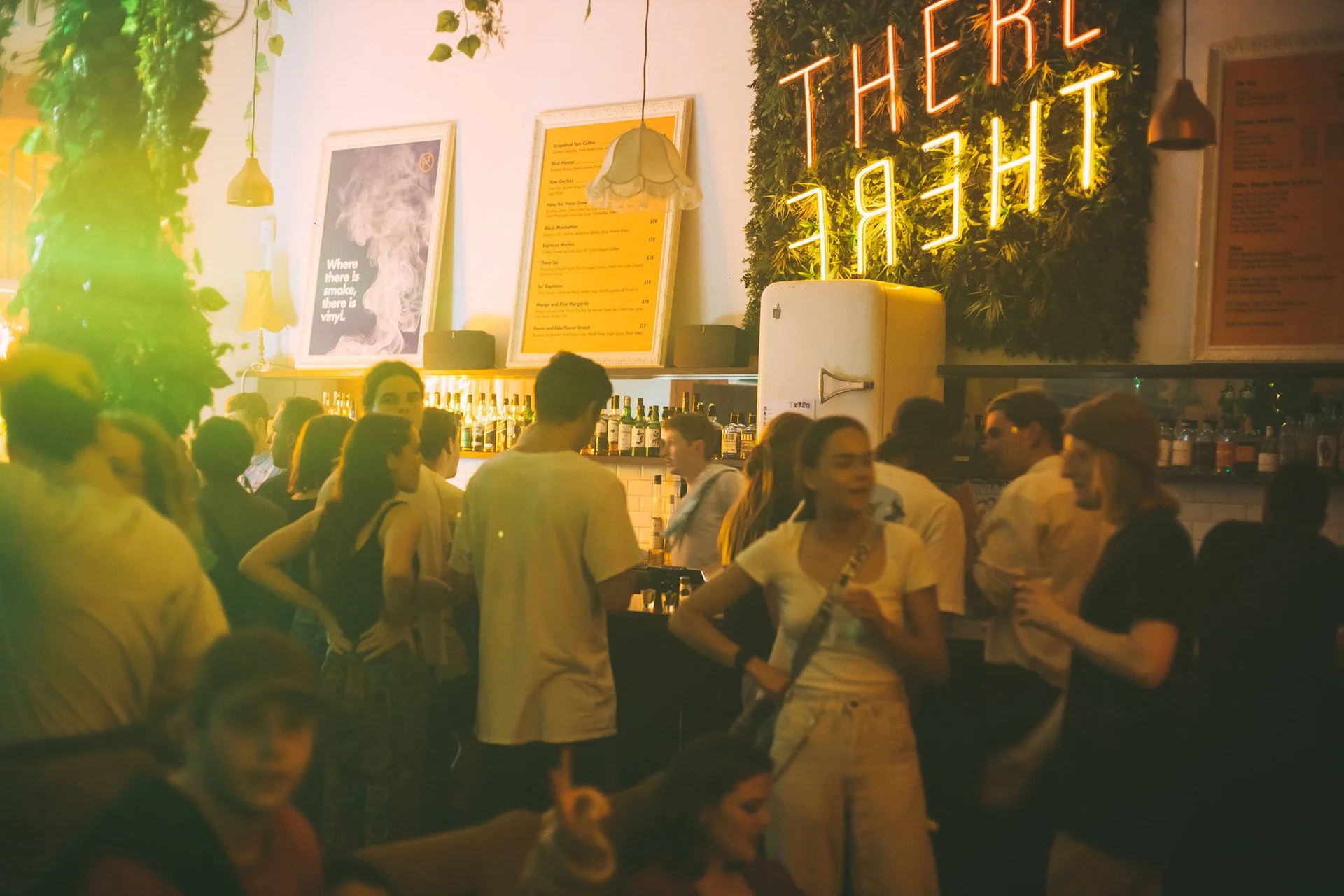 People socializing in a bar with neon sign that reads 'OTHER' and decorative green wall. Dim ambient lighting, bottles on a high shelf, and framed artwork on the wall.